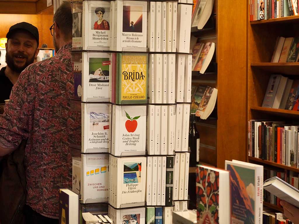 Blick in den Laden: Bücherstapel und Regale, im Vordergrund zwei Besucher